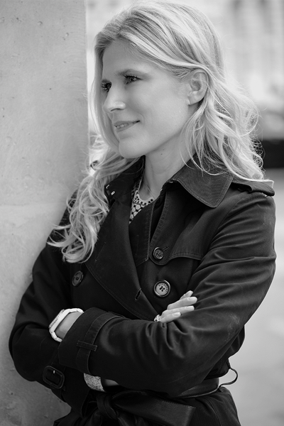 Caroline, leaning against a stone pillar in a Paris street, in a dark trench coat