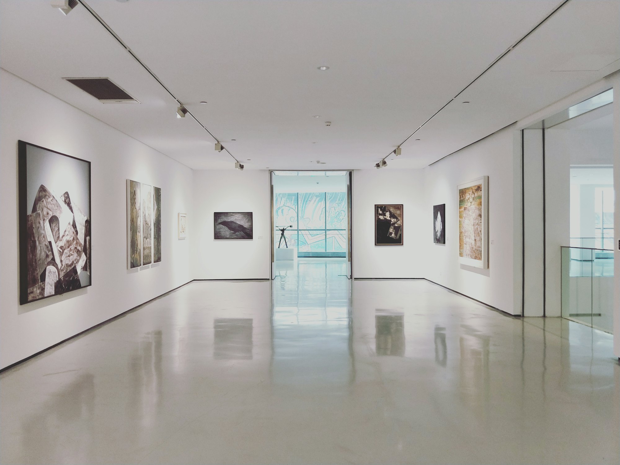 A white gallery corridor under track lighting, works hung along both walls