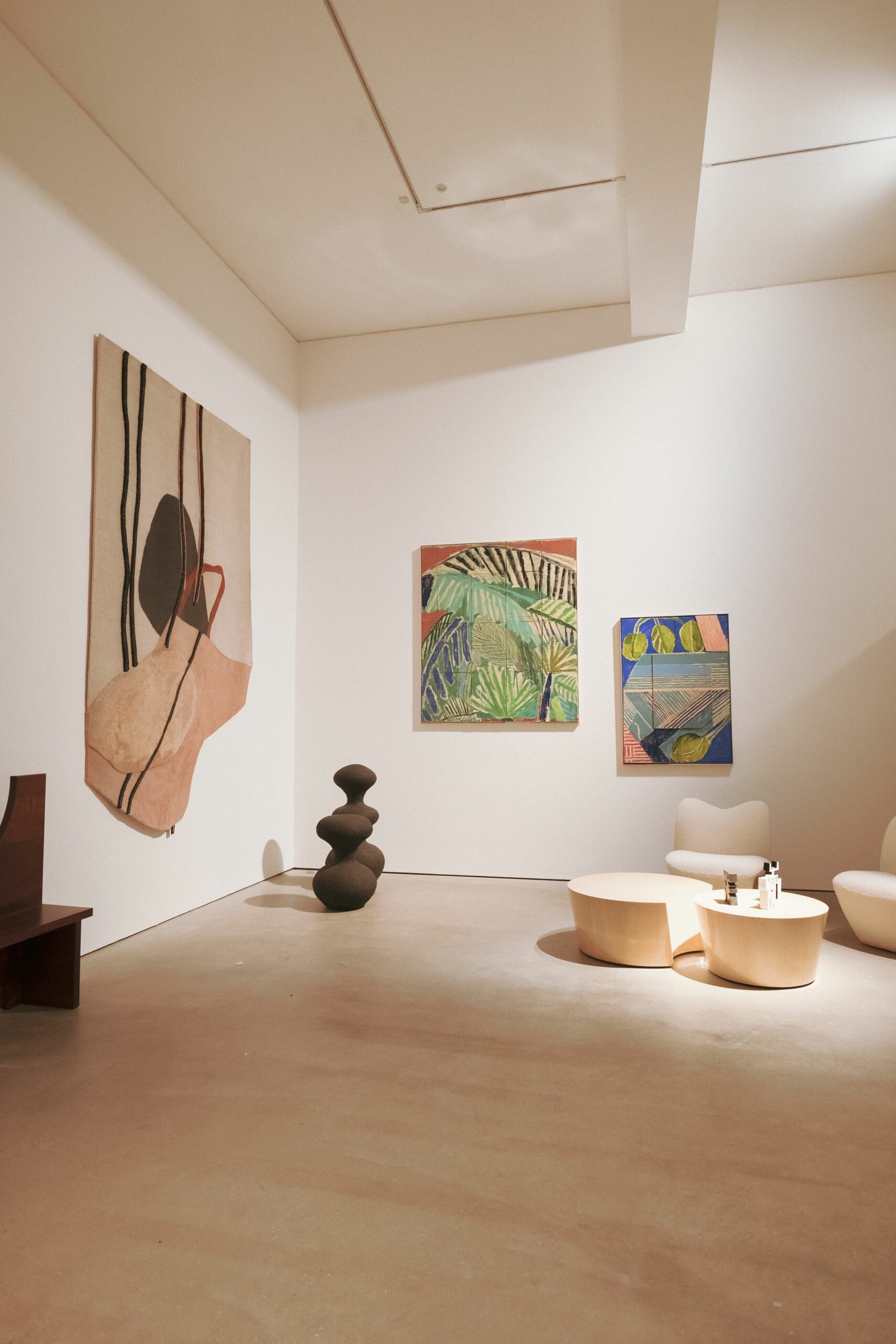 A white gallery room hung with a woven textile and two paintings, a dark ceramic sculpture on the floor and cream seating